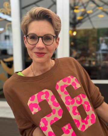 Frau mit kurzen, blonden Haaren und Brille trägt einen braunen Pullover mit großen, pinken Zahlen. Sie lächelt und steht vor einem hellen Hintergrund mit unscharfen Details.