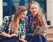 Zwei Frauen sitzen auf einer Treppe und schauen gemeinsam auf ein Tablet. Beide tragen lässige Kleidung und lächeln, während sie sich unterhalten.