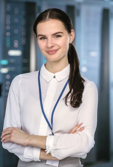 Eine junge Frau mit langen, dunklen Haaren steht in einem Serverraum. Sie trägt eine weiße Bluse und hat einen blauen Ausweis um den Hals. Im Hintergrund sind Serverregale sichtbar.