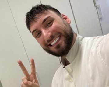 Ein Mann mit Bart und lächelndem Gesicht trägt eine weiße Uniform und zeigt ein Peace-Zeichen mit der Hand.