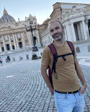 Ein Mann mit Bart steht auf dem Petersplatz in Rom, vor der beeindruckenden Fassade der Peterskirche. Er trägt ein braunes T-Shirt und eine Umhängetasche, während Menschen im Hintergrund spazieren.