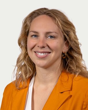 Eine lächelnde Frau mit lockigem, blondem Haar trägt ein orangefarbenes Blazer. Der Hintergrund ist neutral und hell.