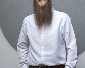 Ein Mann mit einer langen, gepflegten Bart trägt ein weißes Hemd und schwarze Hose. Er steht mit einer Hand in der Hosentasche und lächelt, vor einem grauen Hintergrund.
