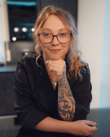 Eine Person mit Brille und tätowiertem Arm lächelt, während sie auf einem Tisch in einer modernen Küche sitzt.