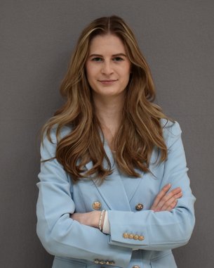 Eine Frau mit langen, lockigen Haaren steht mit verschränkten Armen vor einer grauen Wand. Sie trägt einen hellblauen Blazer und schaut direkt in die Kamera.