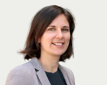 Eine Frau mit schulterlangen, braunen Haaren und einem grauen Blazer lächelt freundlich in die Kamera.