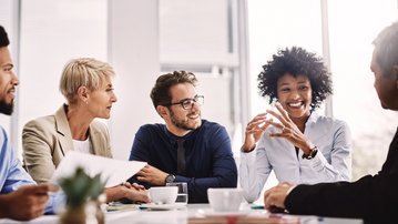 Fünf Personen sitzen an einem Tisch und diskutieren. Eine Frau mit lockigem Haar spricht, während die anderen aufmerksam zuhören. Auf dem Tisch stehen Kaffeetassen und Unterlagen.