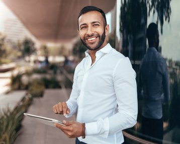 Ein Mann in einem weißen Hemd steht draußen und hält ein Tablet in der Hand. Er lächelt und blickt in die Kamera, während er vor einer modernen Gebäudeoberfläche steht.