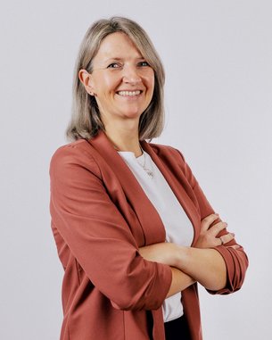 Eine Frau mit schulterlangen, blonden Haaren trägt einen roten Blazer und ein weißes Oberteil. Sie steht mit verschränkten Armen und lächelt.
