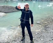 Eine Person in Outdoor-Bekleidung steht am Ufer eines Gletschersees und hebt eine Hand zum Gruß. Im Hintergrund sind Eisblöcke und felsige Ufer zu sehen.
