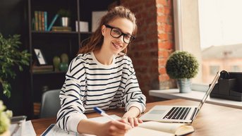 Eine junge Frau mit Brille sitzt an einem Tisch, schreibt in ein Notizbuch und lächelt. Neben ihr steht ein Laptop und einige Unterlagen sind auf dem Tisch verteilt. Im Hintergrund sind Pflanzen und Bücherregale sichtbar.