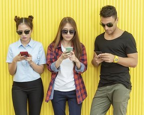 drei junge Menschen mit Handy in der Hand und Sonnenbrille vor Wand stehend und Blick auf die Handys