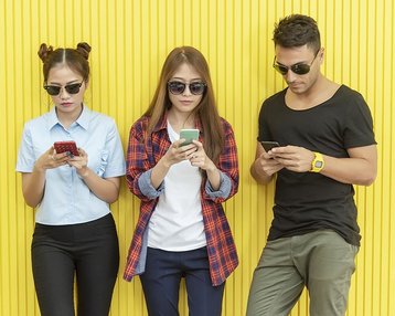 drei junge Menschen mit Handy in der Hand und Sonnenbrille vor Wand stehend und Blick auf die Handys