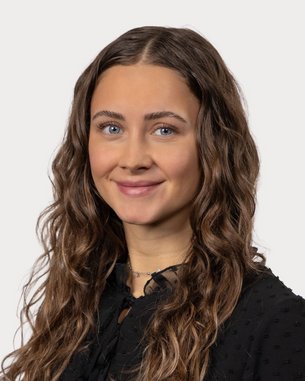 Eine junge Frau mit lockigem, braunem Haar und blauen Augen trägt ein schwarzes Oberteil und lächelt freundlich in die Kamera.