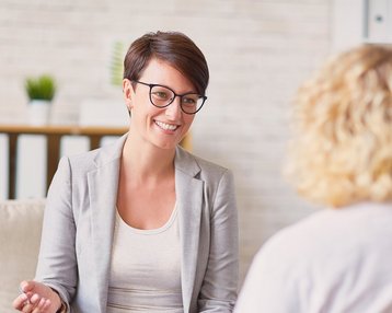 Eine lächelnde Frau mit Brille in einem grauen Blazer sitzt auf einem Sofa und spricht mit einer anderen Person.