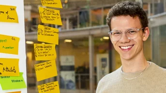 Ein junger Mann mit Brille lächelt, während er an einem Tisch steht, umgeben von bunten Haftnotizen an einer Wand.