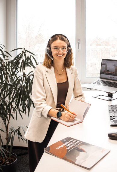 Eine Frau mit Headset sitzt an einem Schreibtisch und arbeitet am Computer, während sie in einem modernen Büro umgeben von Pflanzen ist.