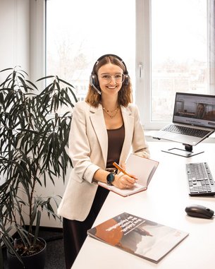 Eine Frau mit Headset sitzt an einem Schreibtisch und arbeitet am Computer, während sie in einem modernen Büro umgeben von Pflanzen ist.