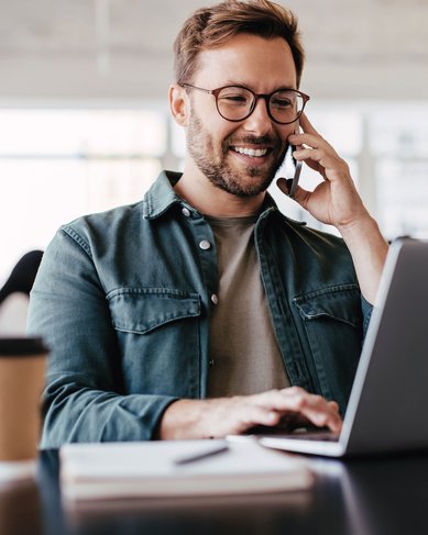 Ein Mann mit Brille sitzt an einem Tisch, spricht am Telefon und tippt auf einem Laptop. Neben ihm steht eine Kaffeetasse und ein Notizbuch liegt auf dem Tisch.
