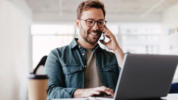 Ein Mann mit Brille sitzt an einem Tisch, spricht am Telefon und tippt auf einem Laptop. Neben ihm steht eine Kaffeetasse und ein Notizbuch liegt auf dem Tisch.