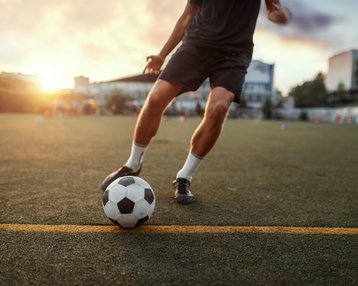 Ein Fußballspieler tritt einen Ball auf einem Rasenplatz bei Sonnenuntergang. Die Szene zeigt den Spieler in Bewegung, während er sich auf das Spiel konzentriert.