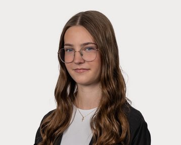 Eine Person mit langen, lockigen Haaren und Brille, die in einem neutralen Hintergrund steht. Sie trägt ein schwarzes Oberteil und hat einen neutralen Gesichtsausdruck.