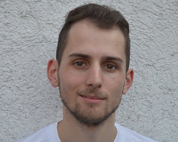 Ein junger Mann mit kurzen Haaren trägt ein weißes T-Shirt mit einem schwarzen Nike-Logo. Er steht vor einer strukturierten, hellen Wand und blickt direkt in die Kamera.