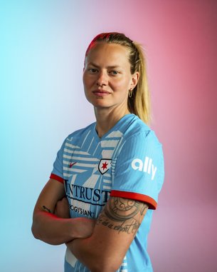 Eine sportliche Frau mit langen blonden Haaren trägt ein hellblaues Fußballtrikot mit roten Akzenten. Sie steht mit verschränkten Armen vor einem farbigen Hintergrund in Blau und Rosa.