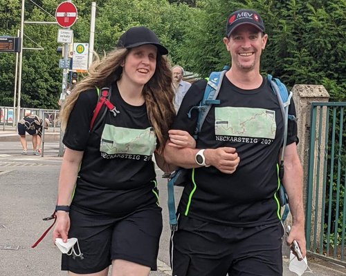 Ein Paar in sportlicher Kleidung mit Rucksäcken geht auf einem Gehweg. Beide tragen T-Shirts mit dem Aufdruck "Neckarsteig 2022". Im Hintergrund sind weitere Personen und Verkehrsschilder sichtbar.