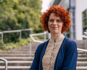 Eine Frau mit roten, lockigen Haaren steht lächelnd vor einer Treppe, die von Bäumen umgeben ist. Sie trägt ein helles Oberteil und eine blaue Jacke.