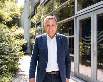 Ein Mann in einem blauen Blazer und weißem Hemd steht lächelnd vor einem modernen Gebäude mit großen Fenstern und einer begrünten Umgebung.