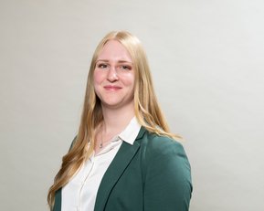 Eine blonde Frau mit langen Haaren trägt ein grünes Blazer und ein weißes Hemd. Sie steht vor einem neutralen Hintergrund und lächelt leicht.