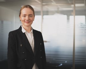 Eine junge Frau in einem schwarzen Blazer steht in einem modernen Büro mit Glaswänden. Sie hat blonde Haare und trägt eine weiße Bluse.