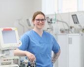 Eine Krankenschwester in blauer Arbeitskleidung steht lächelnd neben medizinischen Geräten in einem modernen Krankenhausumfeld.