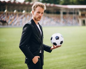 Ein Mann in einem schwarzen Anzug hält einen Fußball in der Hand, während er auf einem Sportfeld steht.