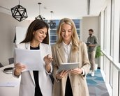 Zwei Geschäftspersonen in Anzügen betrachten gemeinsam ein Tablet in einem modernen Büro. Ein Tisch mit Laptops und Stühlen ist im Hintergrund sichtbar.