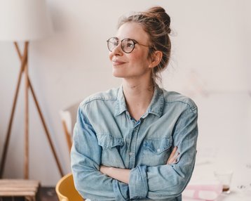 Eine lächelnde Frau mit Brille sitzt entspannt in einem hellen Raum, trägt ein Jeanshemd und hat die Arme verschränkt.