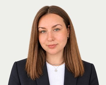 Eine Person mit langen, braunen Haaren und grünen Augen trägt einen schwarzen Blazer und ein weißes Oberteil.