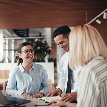 Drei Personen in einem modernen Büro diskutieren und lächeln. Eine Frau mit Brille trägt ein hellblaues Hemd, während die anderen beiden in formeller Kleidung sind. Ein Laptop und Dokumente liegen auf dem Tisch.