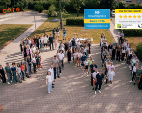 Gruppenszene mit vielen Personen auf einem Platz, die zusammenstehen. Auszeichnungen für eine Fernschule sind im Bild sichtbar.