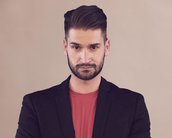 Ein Mann mit Bart trägt einen schwarzen Blazer über einem roten Shirt. Er steht mit verschränkten Armen und hat einen neutralen Gesichtsausdruck. Der Hintergrund ist einfarbig und hell.
