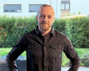 Mann mit kurzem Bart und Glatze, trägt ein schwarzes Hemd, steht mit Händen in der Hüfte vor einer grünen Hecke.