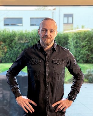 Mann mit kurzem Bart und Glatze, trägt ein schwarzes Hemd, steht mit Händen in der Hüfte vor einer grünen Hecke.