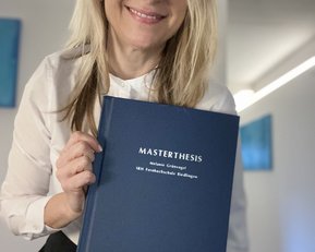 Eine Frau hält stolz ihre Masterarbeit in der Hand. Der Titel "MASTERTHESIS" und ihr Name "Melanie Grunigel" sind auf dem Cover zu sehen. Der Hintergrund ist hell und modern gestaltet.
