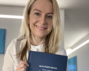 Eine Frau hält stolz ihre Masterarbeit in der Hand. Der Titel "MASTERTHESIS" und ihr Name "Melanie Grunigel" sind auf dem Cover zu sehen. Der Hintergrund ist hell und modern gestaltet.