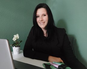 Eine Frau mit langen, dunklen Haaren sitzt an einem Tisch, lächelt und trägt einen schwarzen Blazer. Auf dem Tisch befinden sich ein Laptop, Schreibutensilien und eine kleine Pflanze in einem Topf.