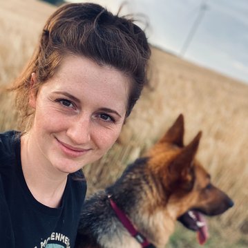 Eine Person mit lockigem Haar lächelt in die Kamera, während ein Hund neben ihr sitzt. Im Hintergrund erstreckt sich ein Feld mit hohem Gras.