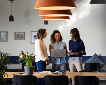 Drei Frauen stehen um einen Tisch in einem modernen Büro und diskutieren. Eine Frau hält einen Laptop, während die anderen zwei auf den Bildschirm schauen. Pflanzen und Möbel sind im Hintergrund sichtbar.