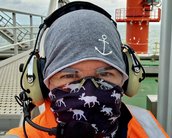 Eine Person trägt einen grauen Kopfschutz und ein schwarzes Tuch mit Tiermotiven. Sie hat Kopfhörer auf und steht auf einer Plattform mit einem roten Kran im Hintergrund.