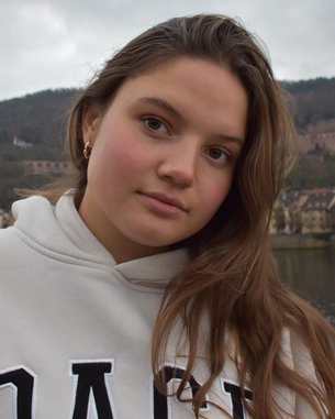 Ein junges Mädchen mit langen, braunen Haaren trägt einen weißen Hoodie. Sie steht an einem Geländer mit Blick auf einen Fluss und eine malerische Stadt im Hintergrund, umgeben von Hügeln.